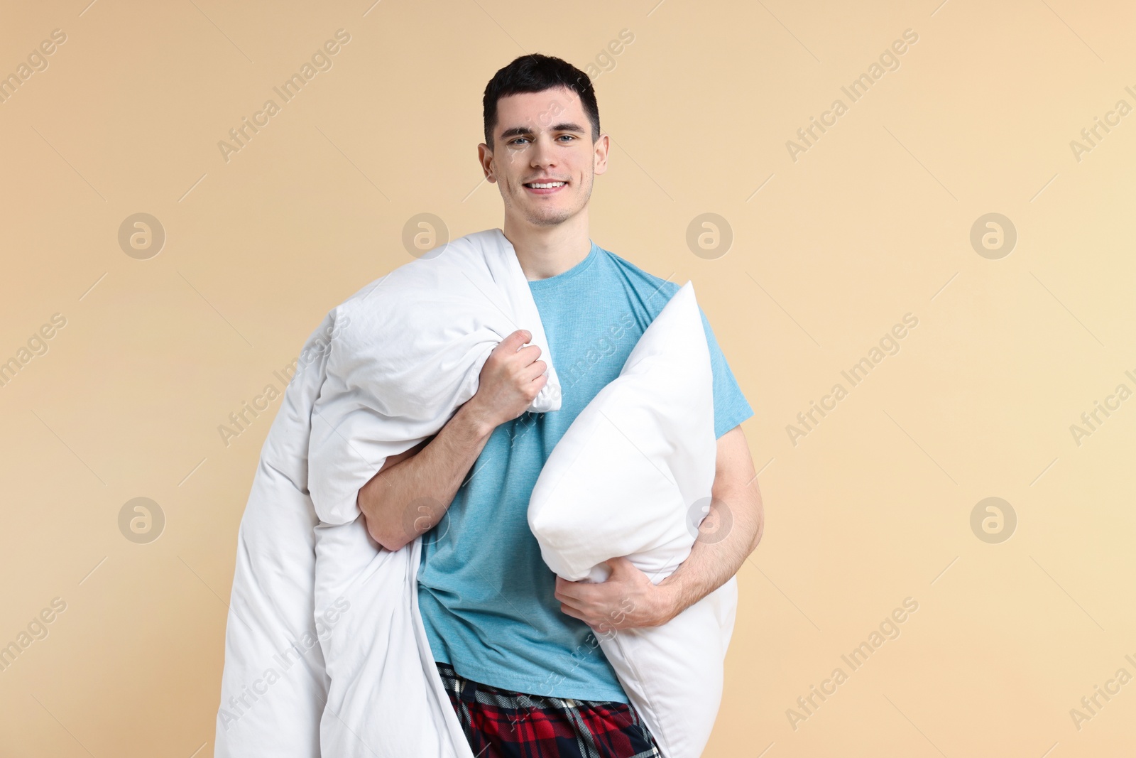 Photo of Happy man in pyjama holding pillow and blanket on beige background