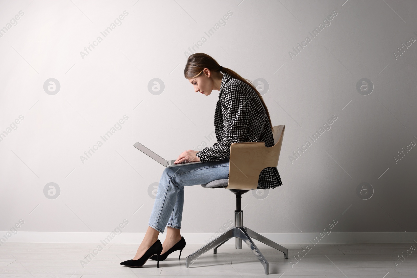Photo of Woman with bad posture using laptop while sitting on chair near light grey wall indoors
