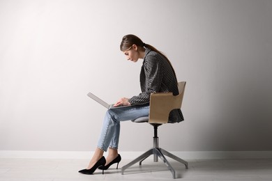 Woman with bad posture using laptop while sitting on chair near light grey wall indoors