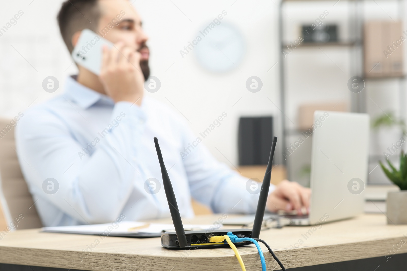 Photo of Man talking on smartphone while working at wooden table indoors, focus on Wi-Fi router