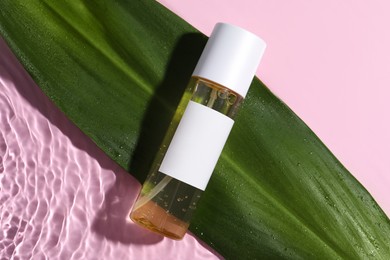 Bottle of cosmetic oil with green leaf in water on pink background, flat lay