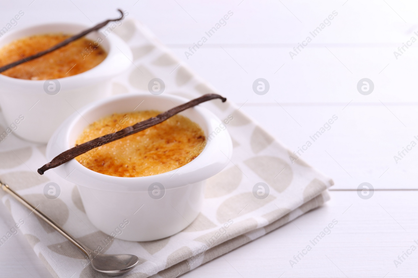 Photo of Delicious creme brulee in bowls, vanilla pods and spoon on white wooden table, closeup