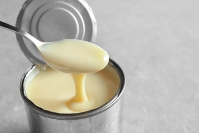 Condensed milk pouring from spoon into tin can on grey background, space for text. Dairy product