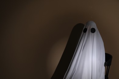 Creepy ghost. Woman covered with sheet sitting on chair against brown background, space for text