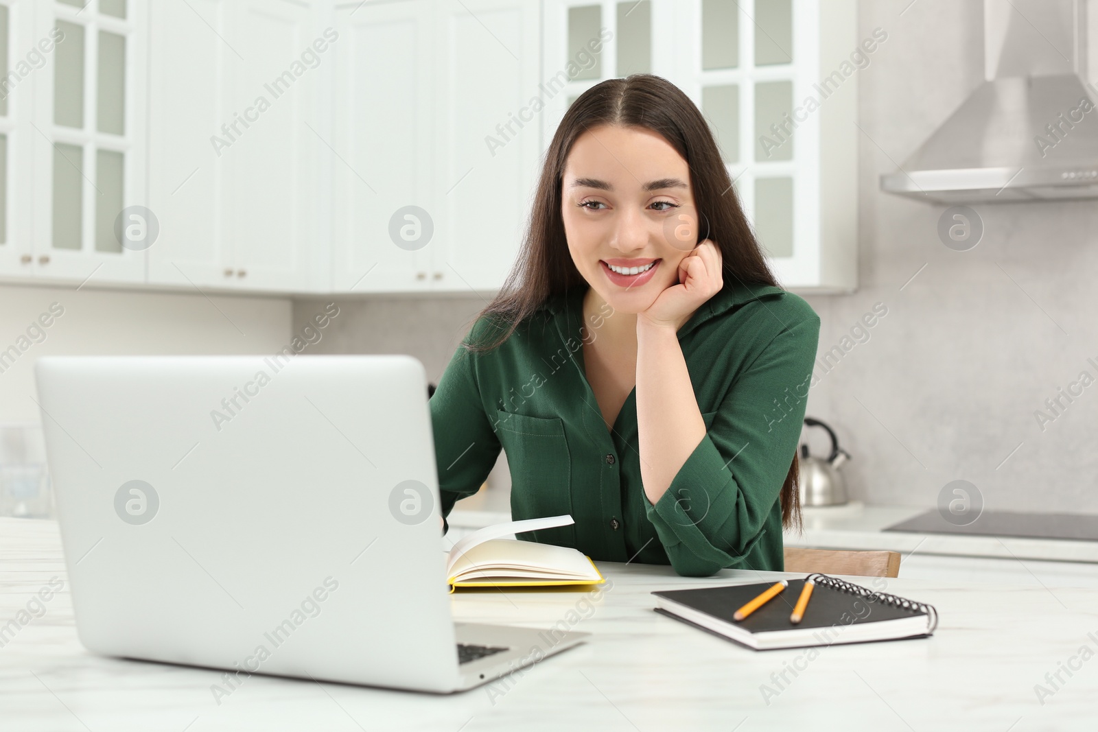 Photo of Home workplace. Happy woman with pen and notebook working on laptop at marble desk in kitchen