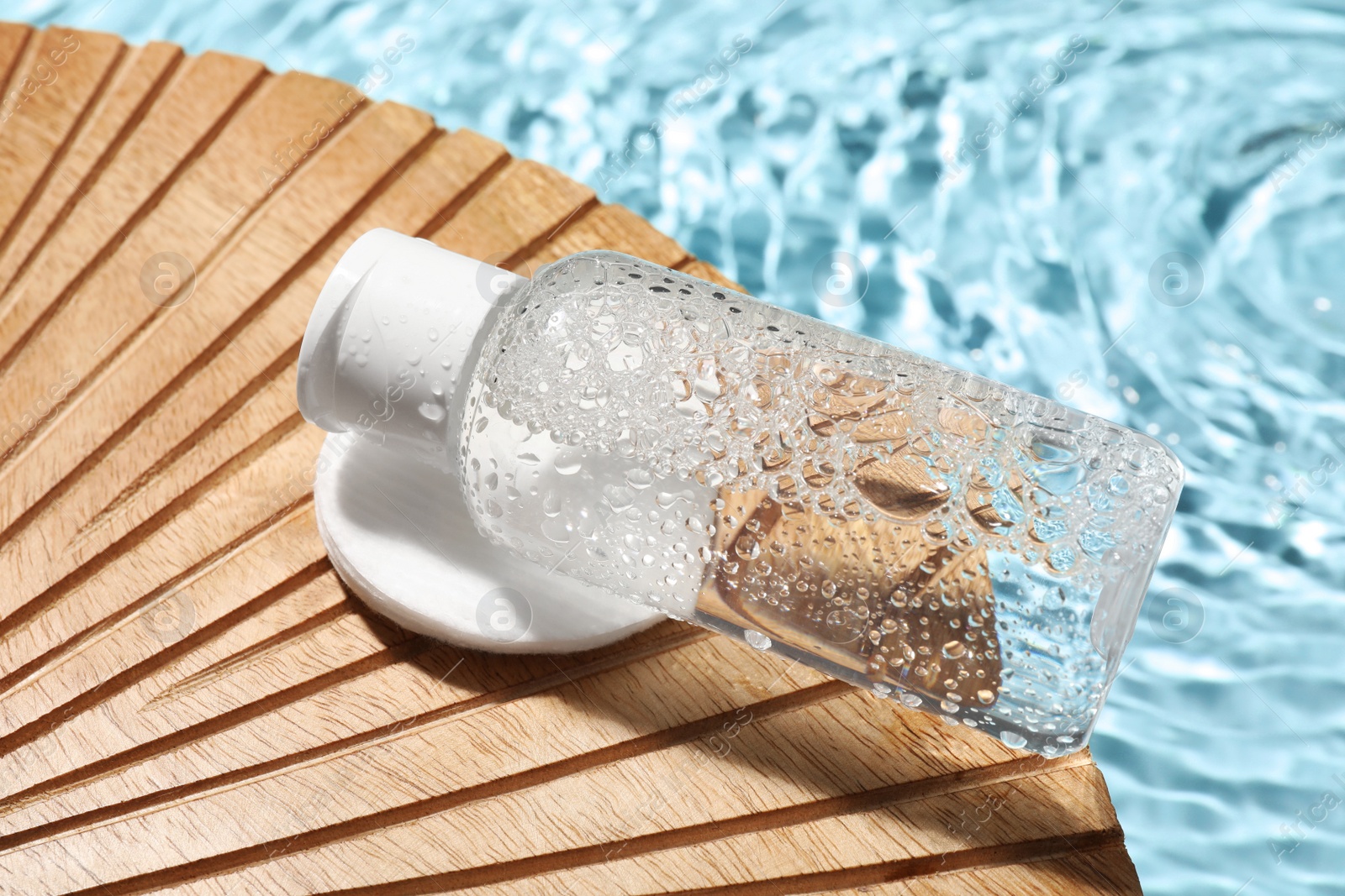 Photo of Wet bottle of micellar water and cotton pad on wooden board, closeup