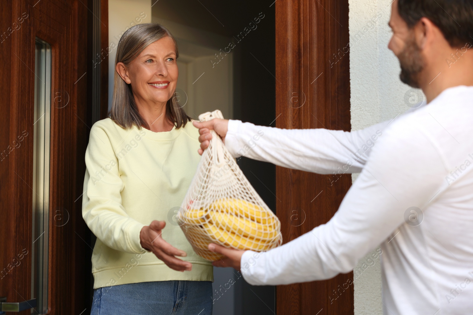 Photo of Helping neighbours. Man with net bag of products visiting senior woman outdoors