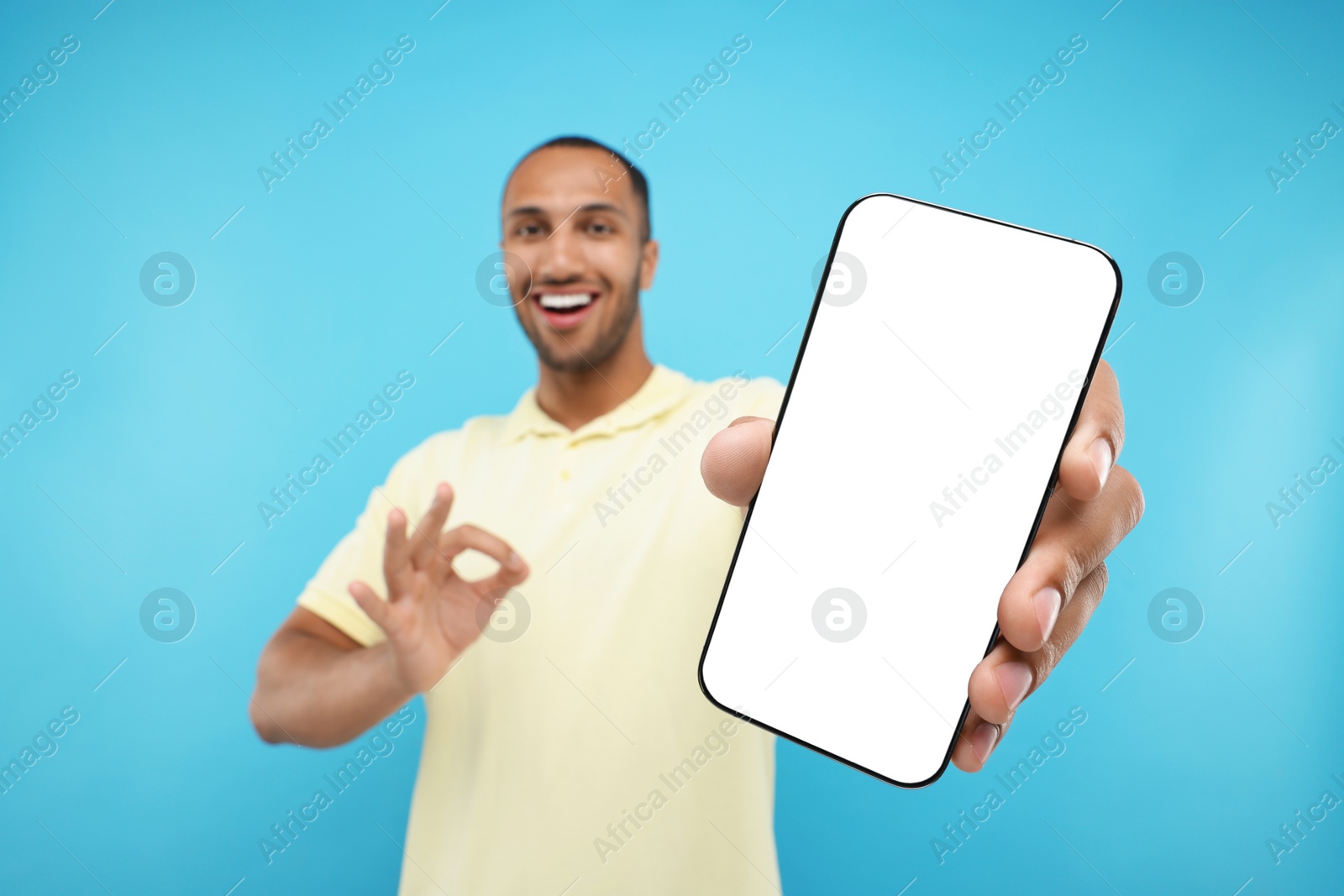 Photo of Young man showing smartphone in hand and OK gesture on light blue background, selective focus. Mockup for design