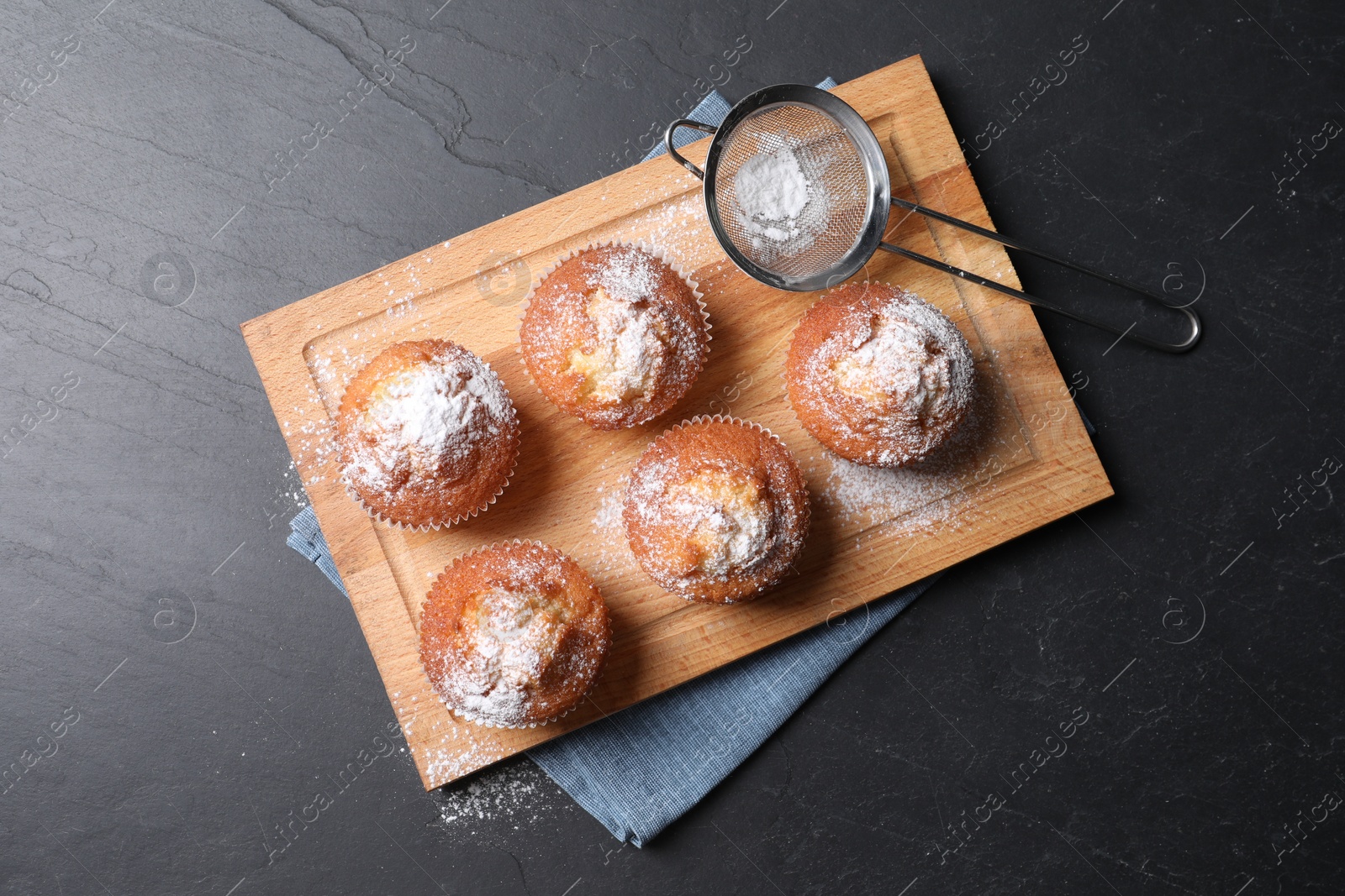 Photo of Delicious sweet muffins and sieve on black textured table, top view