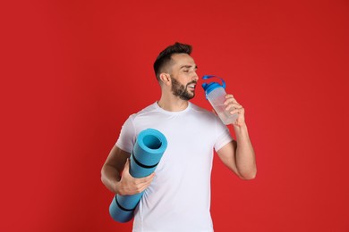 Photo of Handsome man with yoga mat drinking water on red background