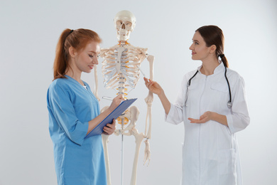 Professional orthopedist with human skeleton model teaching medical student against light background