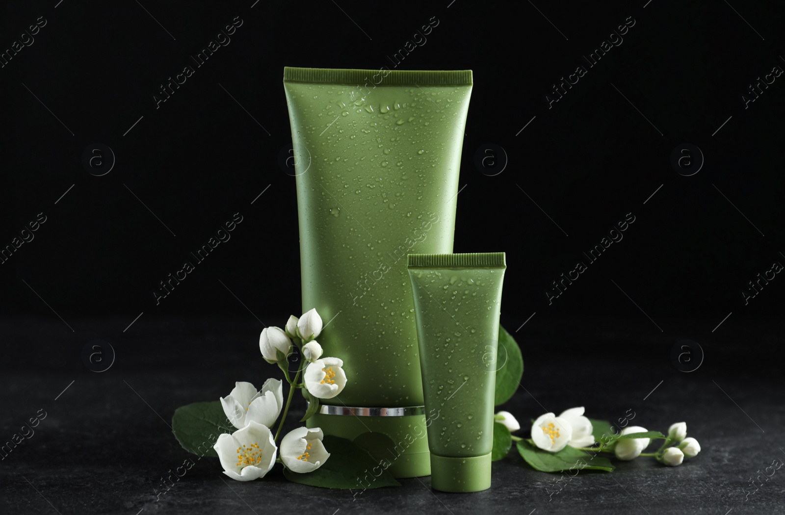 Photo of Tubes of cosmetic products and flowers on grey stone table against black background