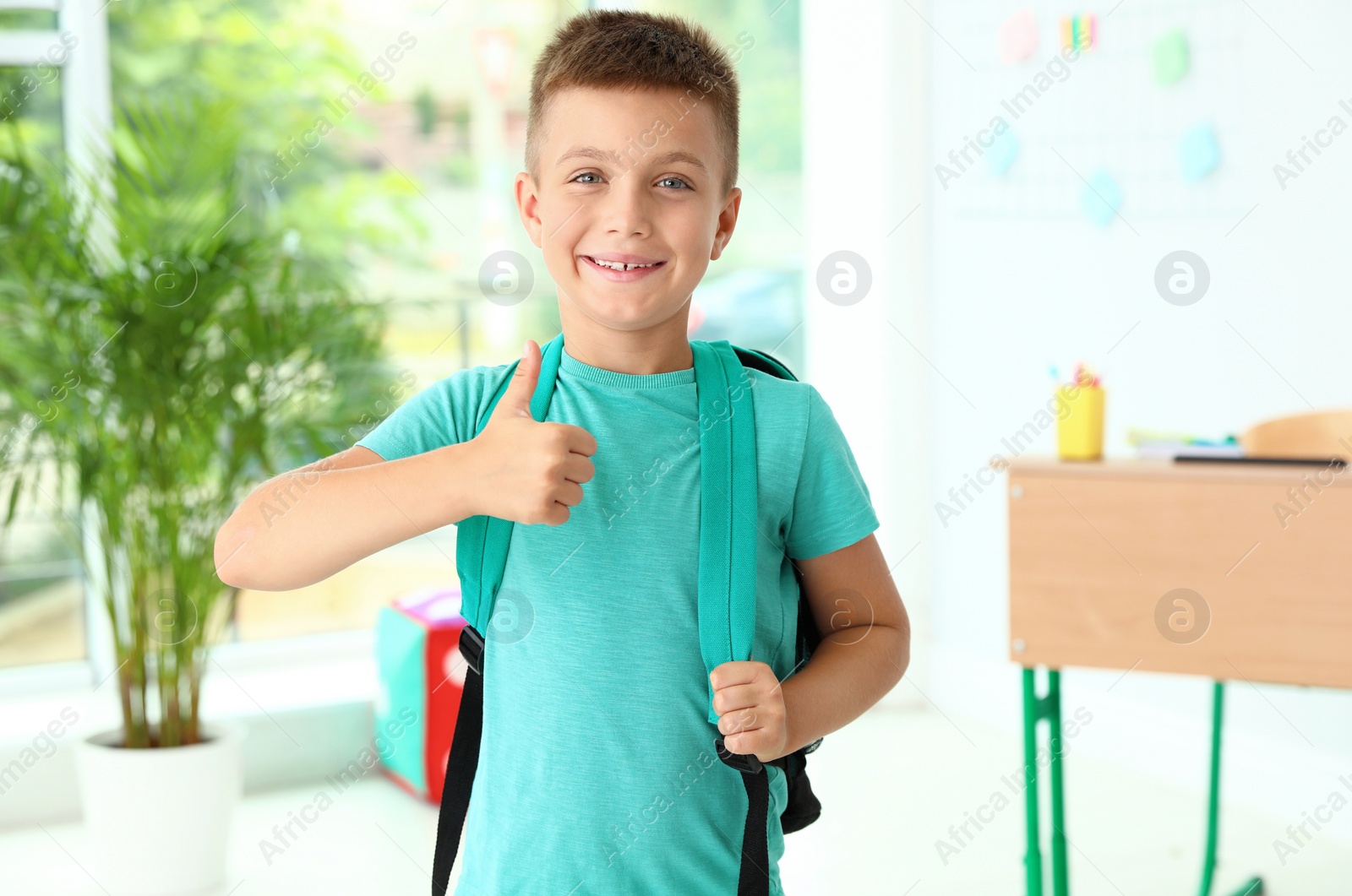 Photo of Cute little boy with backpack showing thumbs-up in classroom at school