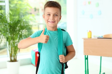 Photo of Cute little boy with backpack showing thumbs-up in classroom at school