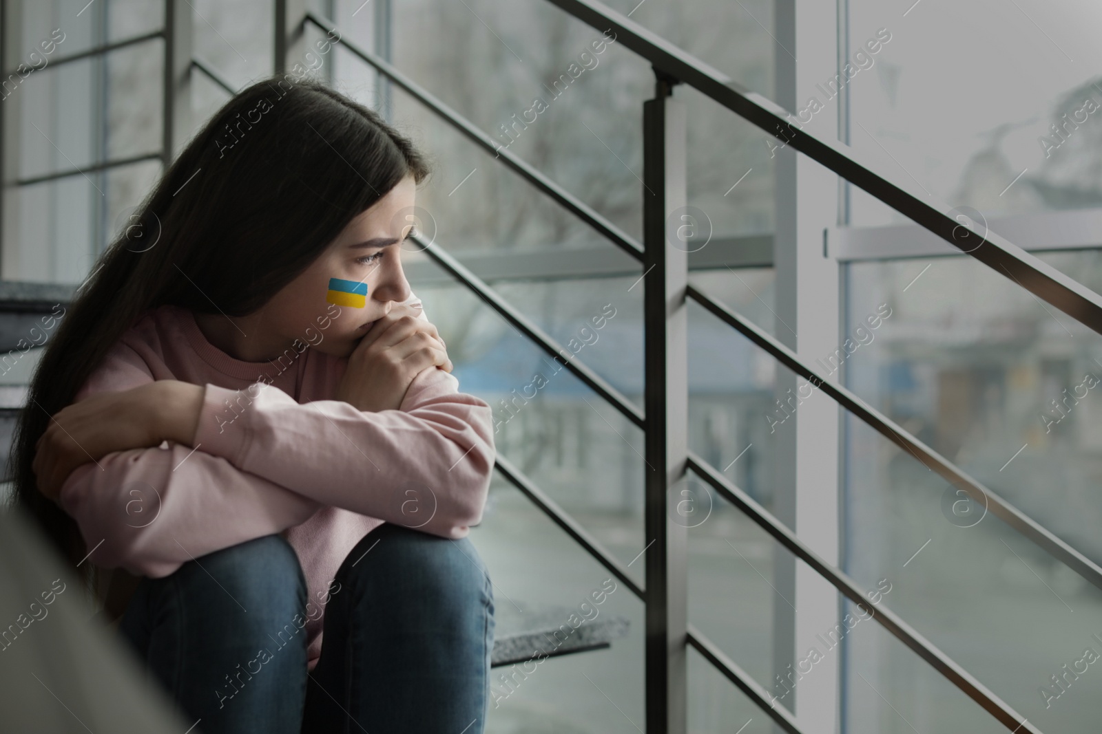 Image of Upset teenage girl with picture of Ukrainian flag on cheek sitting on stairs indoors. Stop war in Ukraine