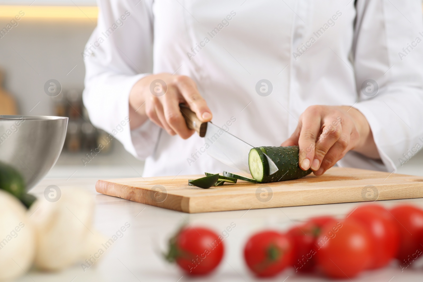 Photo of Professional chef cutting fresh cucumber at white table in kitchen, closeup