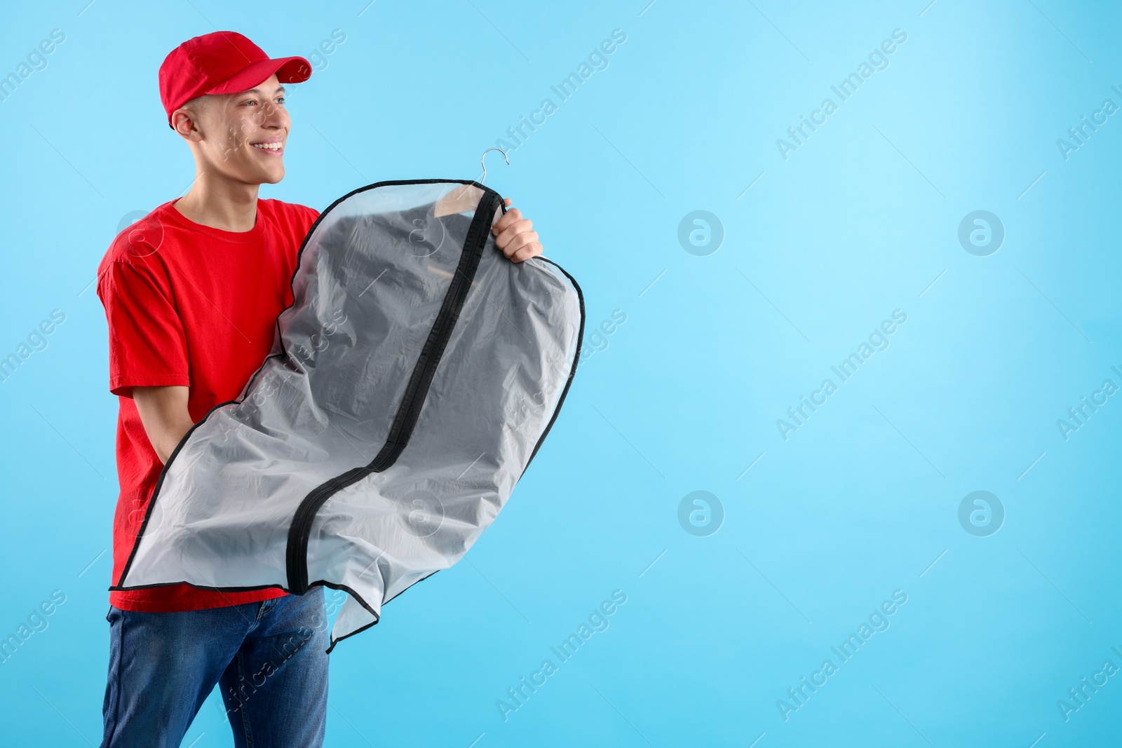 Photo of Dry-cleaning delivery. Happy courier holding garment cover with clothes on light blue background, space for text