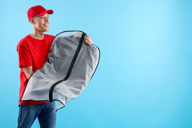 Dry-cleaning delivery. Happy courier holding garment cover with clothes on light blue background, space for text