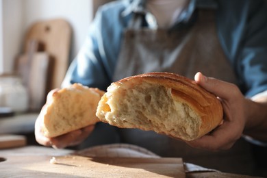 Photo of Man breaking loaf of fresh bread at wooden table indoors, closeup