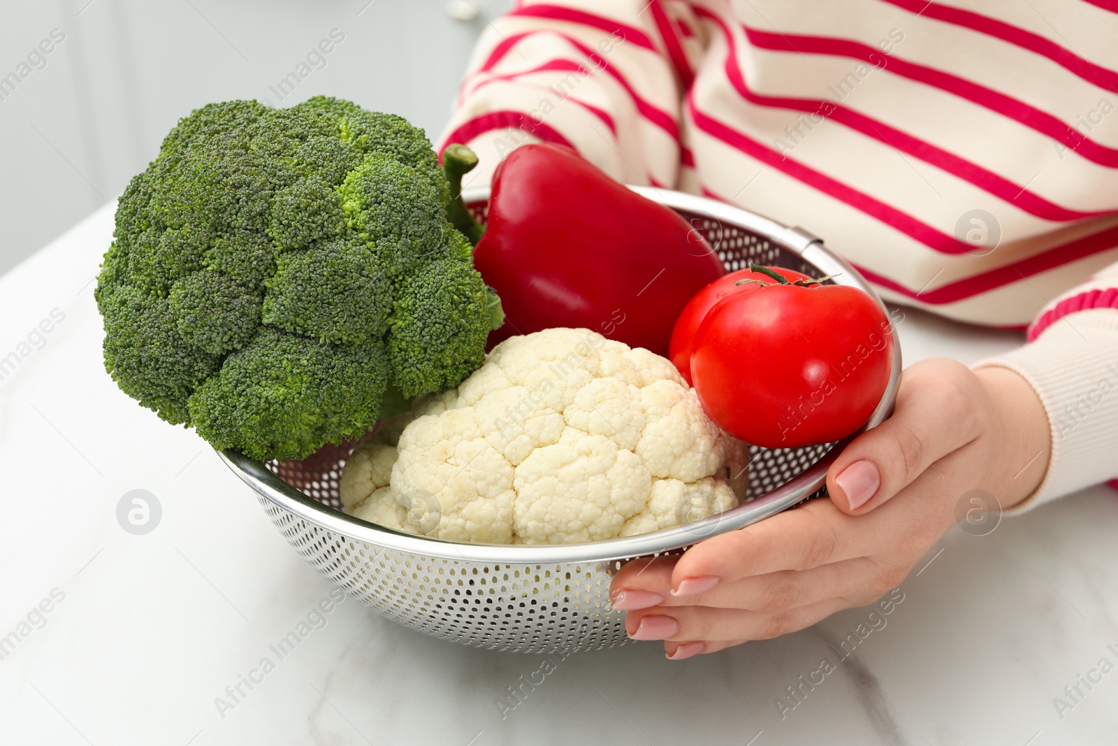 Photo of Woman holding colander with fresh vegetables at white marble table, closeup