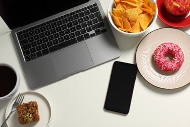 Photo of Bad eating habits at workplace. Laptop, smartphone and different snacks on white table, flat lay