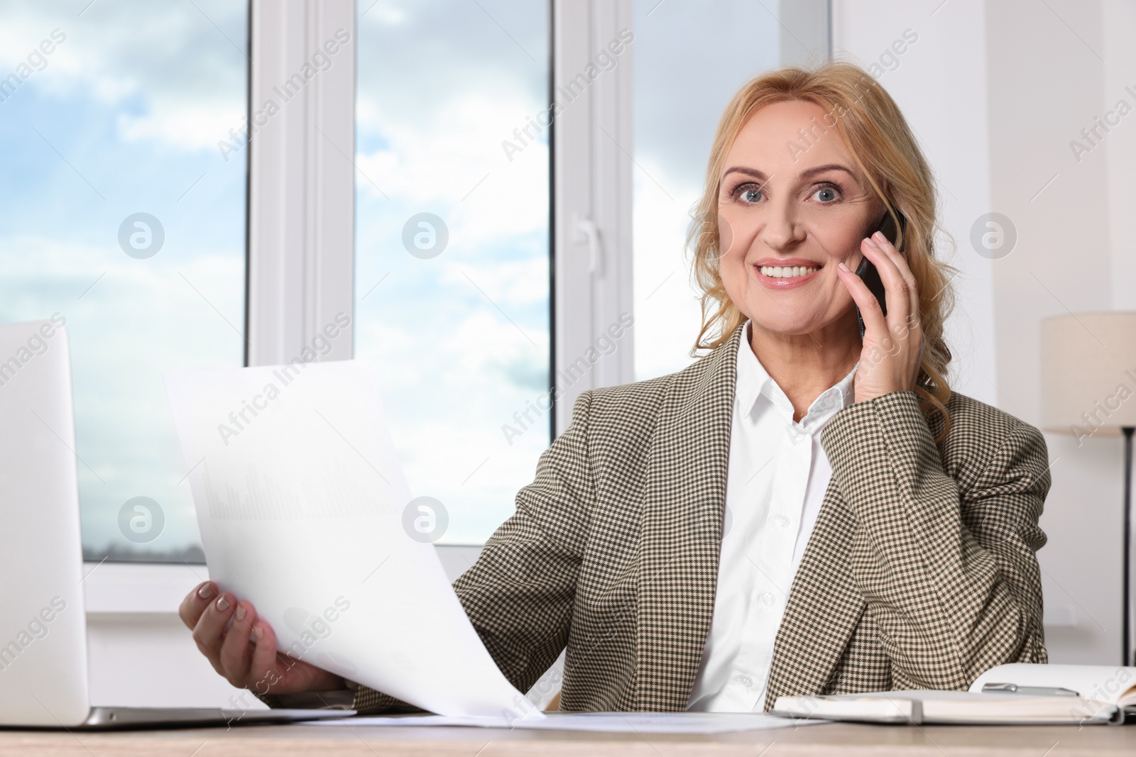 Photo of Lady boss with papers talking on smartphone at desk in office. Successful businesswoman