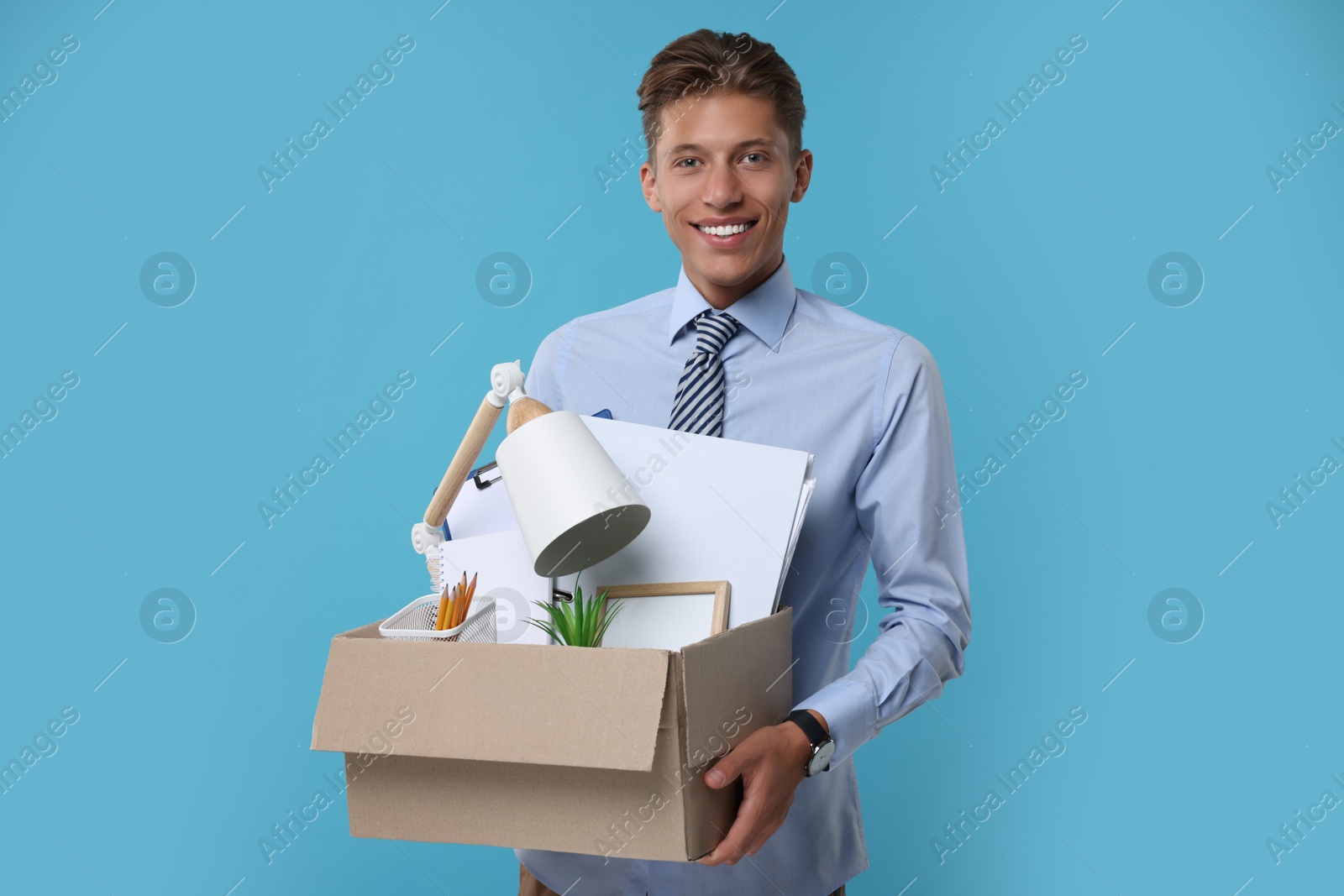 Photo of Happy unemployed young man with box of personal office belongings on light blue background