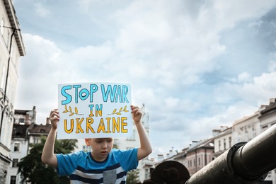 Photo of Boy holding poster in colors of national flag with words Stop War In Ukraine near broken military tank on city street, closeup