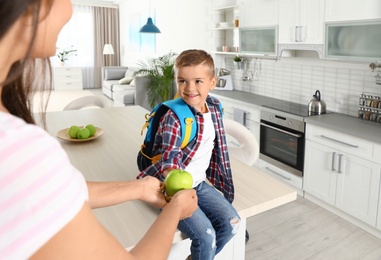 Happy mother giving apple to little child's with school bag in kitchen