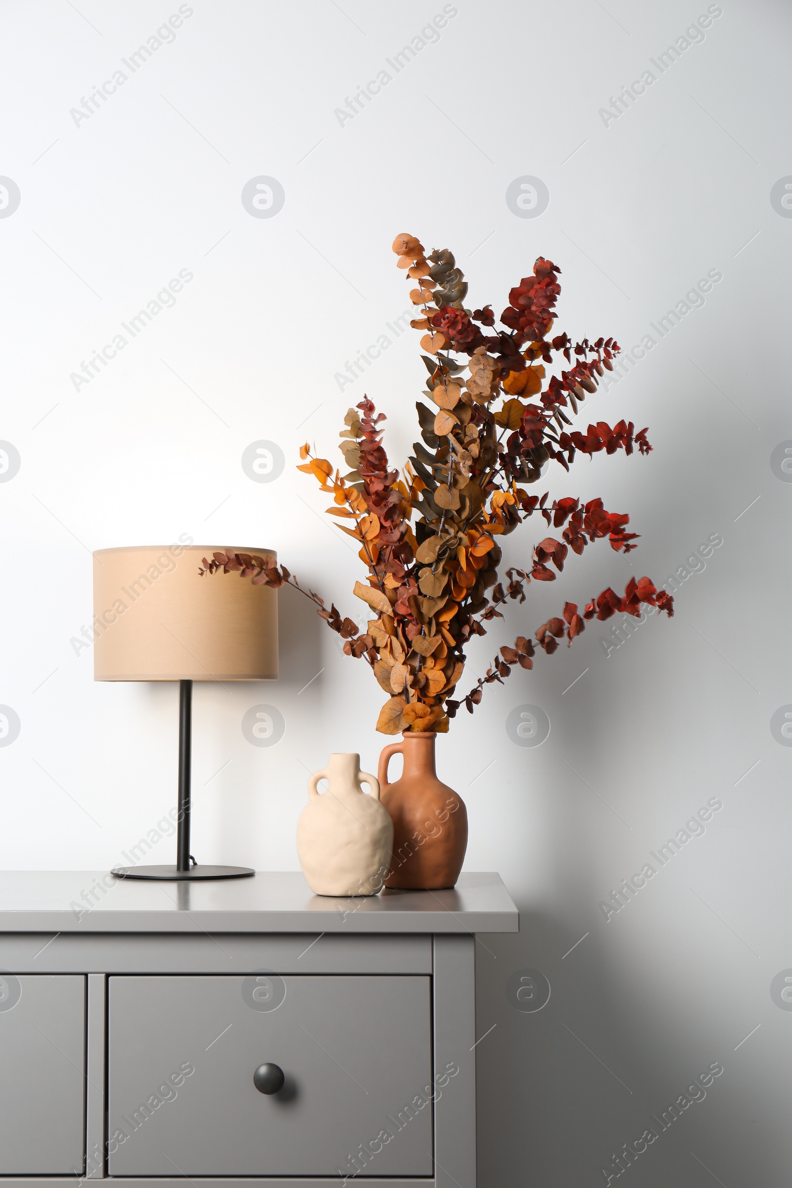 Photo of Stylish vases, dried eucalyptus branches and table lamp on chest of drawers near white wall indoors. Interior design