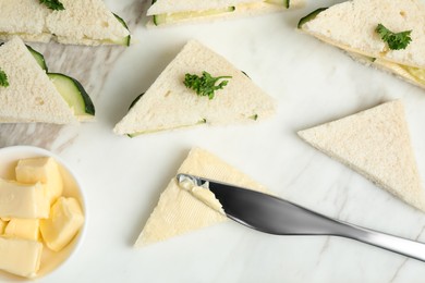 Photo of Spreading butter on tasty sandwiches with cucumber and parsley on white marble table, flat lay