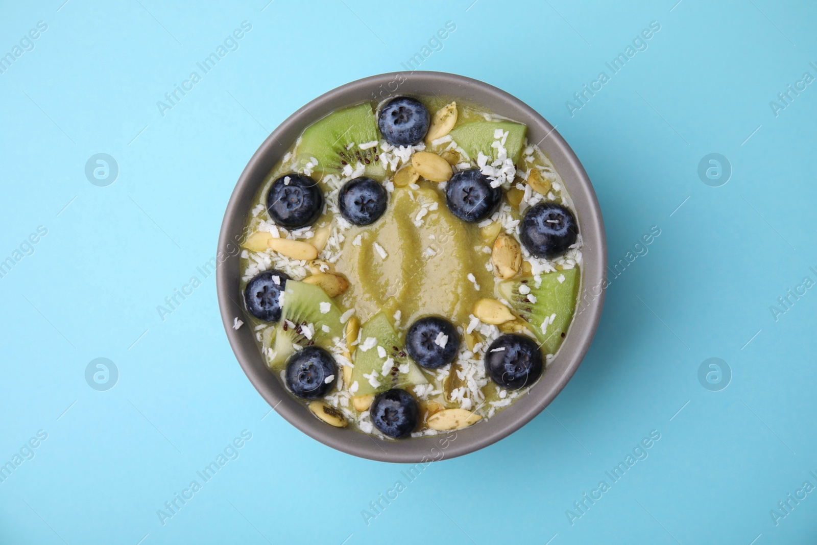 Photo of Bowl of delicious fruit smoothie with fresh blueberries, kiwi slices and coconut flakes on light blue background, top view