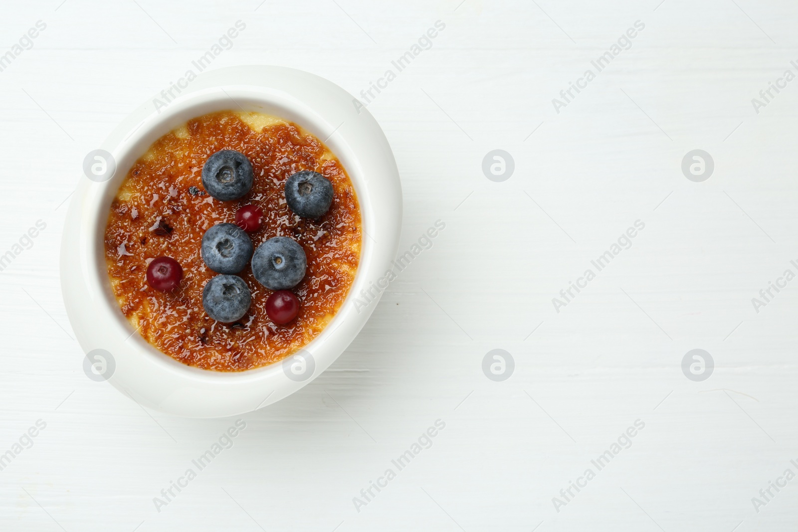 Photo of Delicious creme brulee with berries in bowl on white wooden table, top view. Space for text