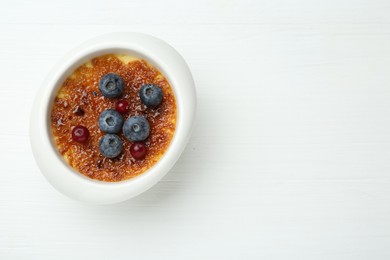 Photo of Delicious creme brulee with berries in bowl on white wooden table, top view. Space for text