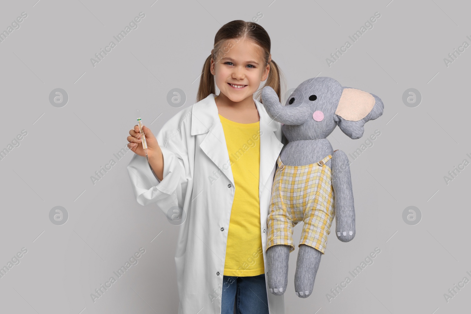 Photo of Little girl in medical uniform with thermometer and toy elephant on light grey background