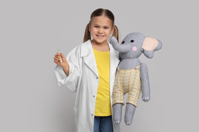 Little girl in medical uniform with thermometer and toy elephant on light grey background