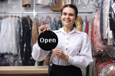 Photo of Dry-cleaning service. Happy worker holding Open sign indoors