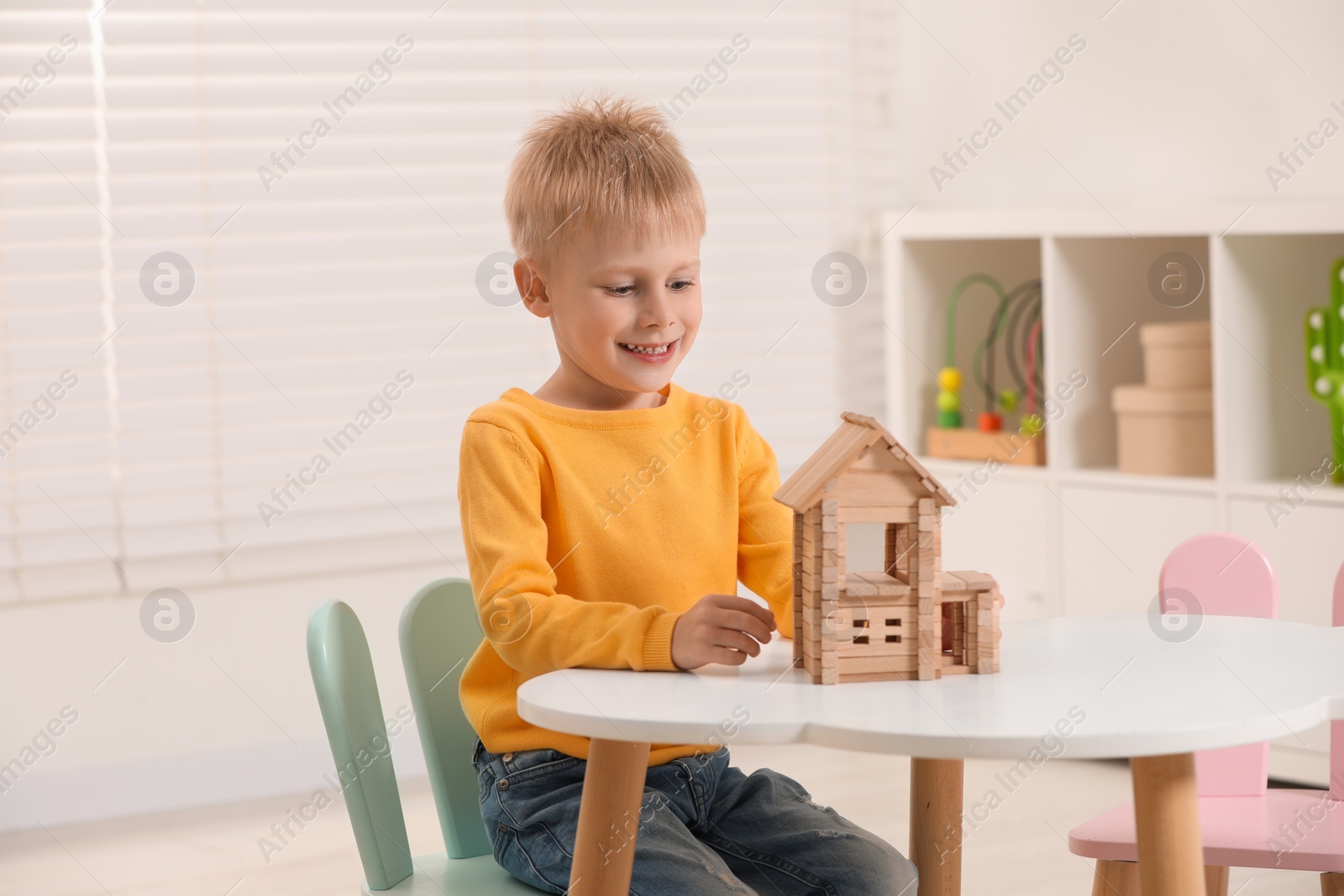 Photo of Cute little boy playing with wooden house at white table indoors. Child's toy