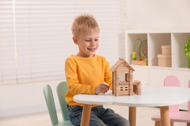 Cute little boy playing with wooden house at white table indoors. Child's toy