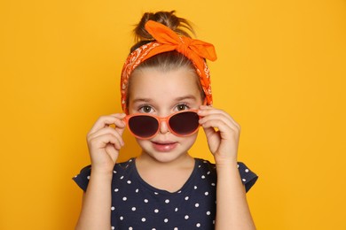 Photo of Cute little girl wearing stylish bandana and sunglasses on orange background