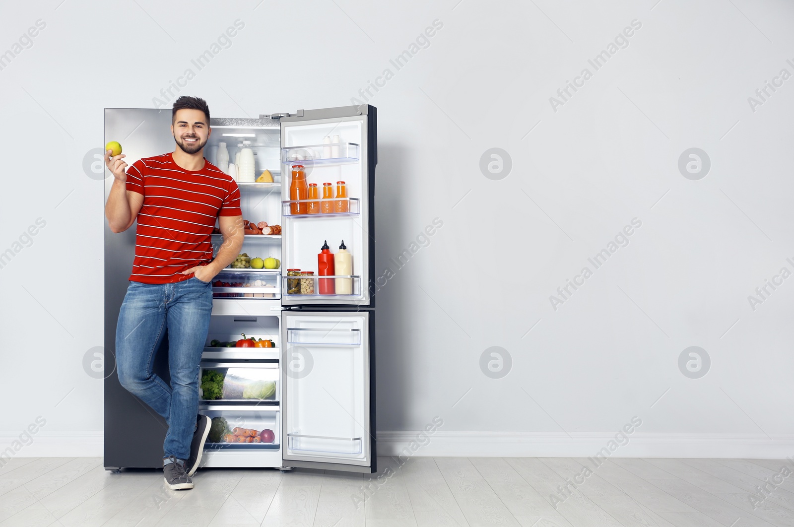 Photo of Happy young man with apple near open refrigerator indoors, space for text