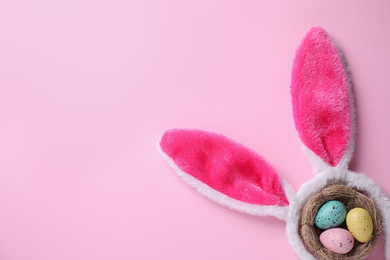 Headband with Easter bunny ears and dyed eggs in nest on pink background, flat lay. Space for text