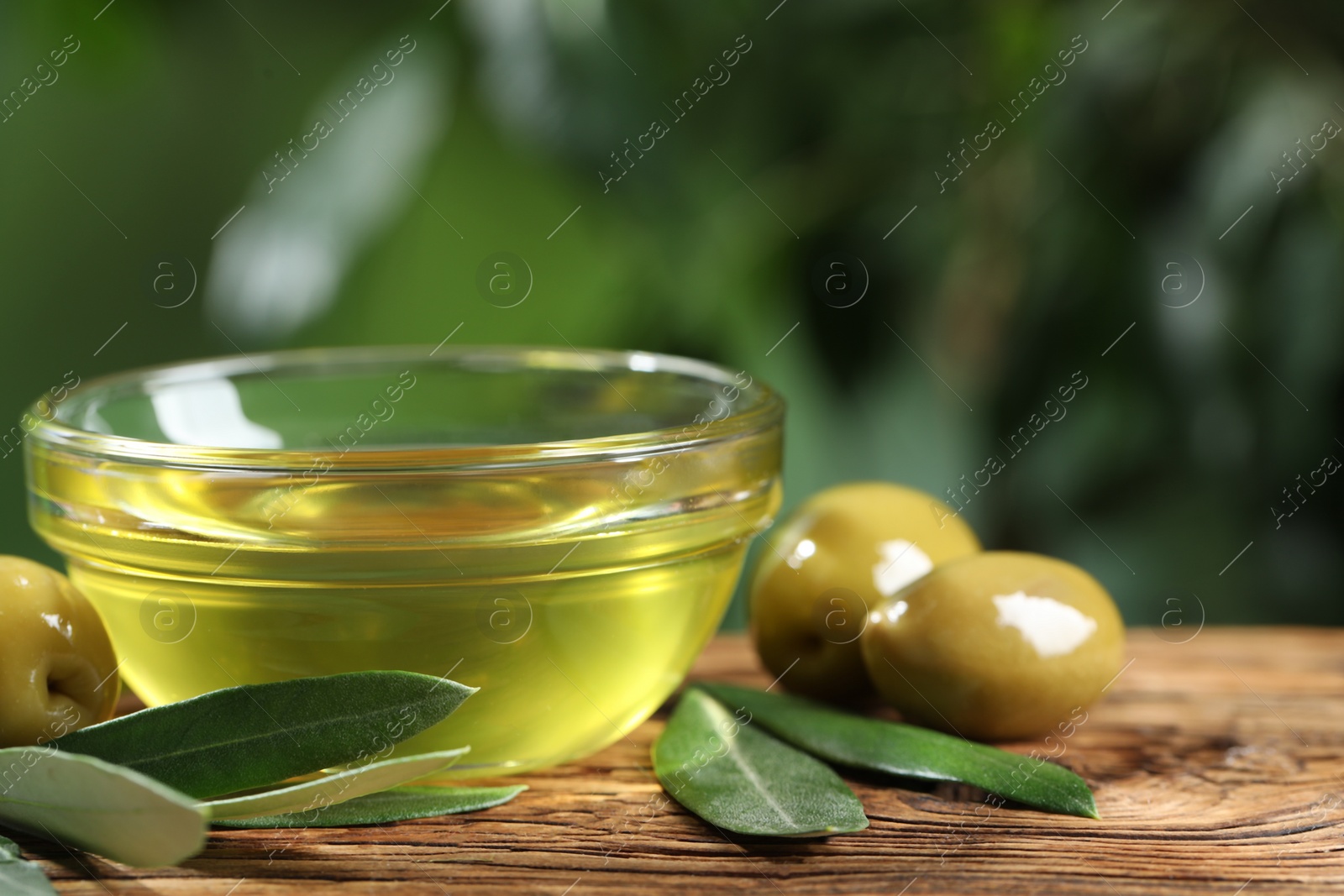 Photo of Bowl with cooking oil, olives and green leaves on wooden table, closeup