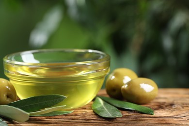 Bowl with cooking oil, olives and green leaves on wooden table, closeup