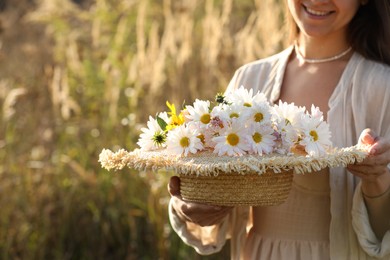 Photo of Woman holding straw hat with beautiful wild flowers outdoors, closeup. Space for text