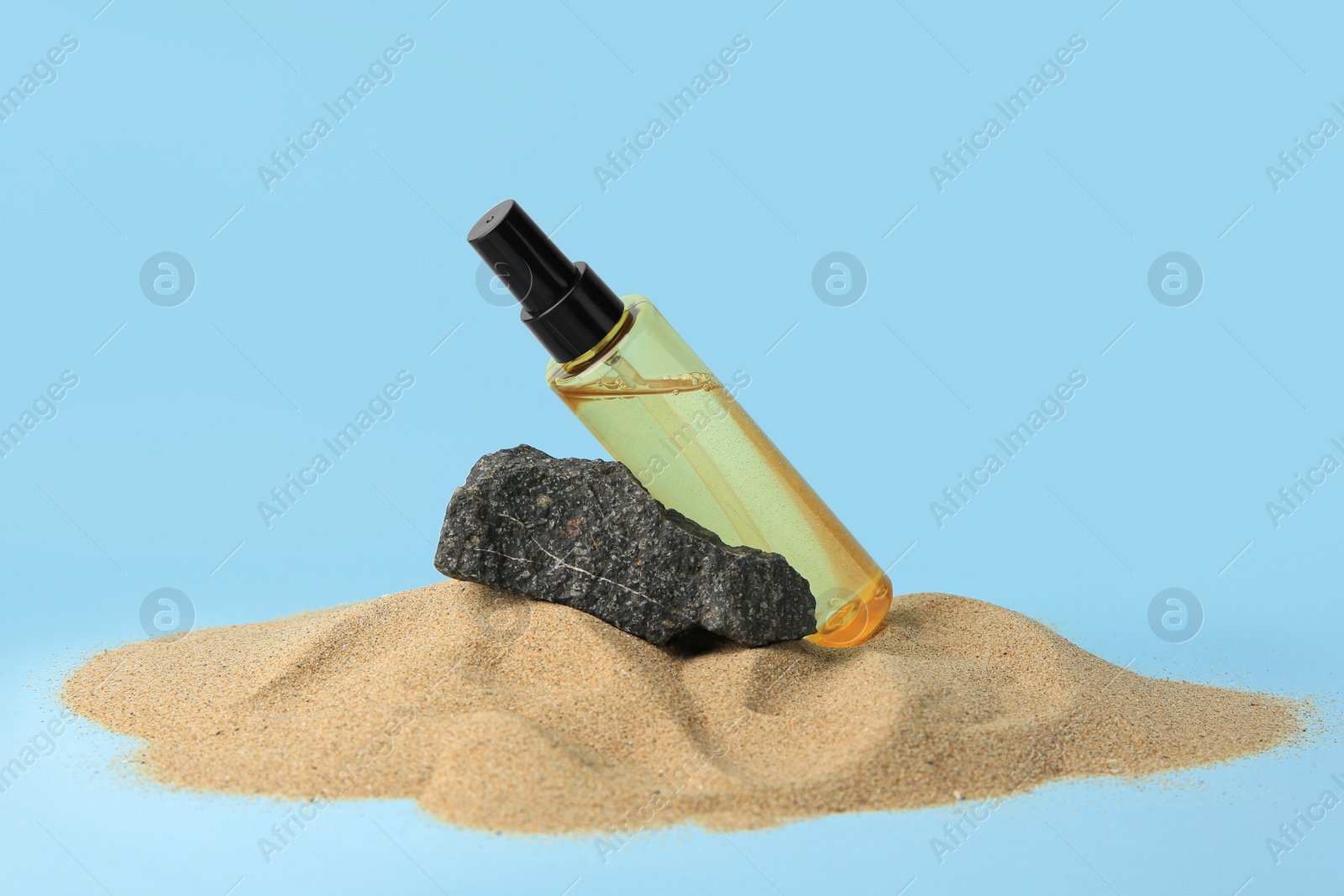 Photo of Bottle with serum and stone on sand against light blue background. Cosmetic product