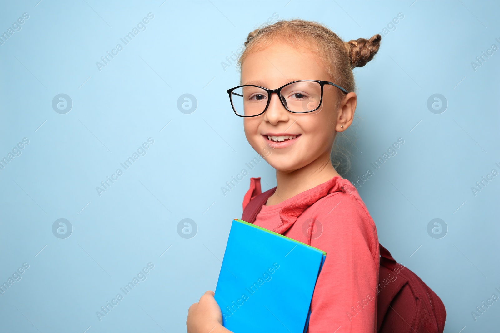 Photo of Cute little girl with glasses, backpack and textbook on light blue background. Space for text