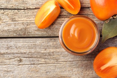 Photo of Delicious persimmon jam in glass jar and fresh fruits on wooden table, flat lay. Space for text