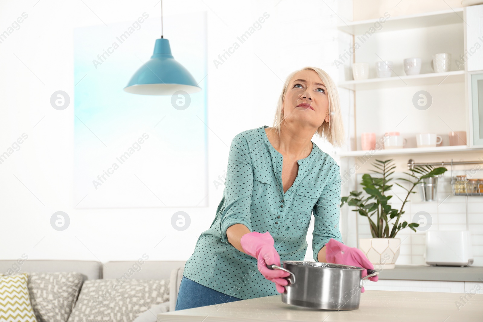 Photo of Worried mature woman collecting water leakage from ceiling at home. Time to call plumber