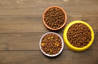 Photo of Dry food in pet bowls on wooden background, flat lay. Space for text